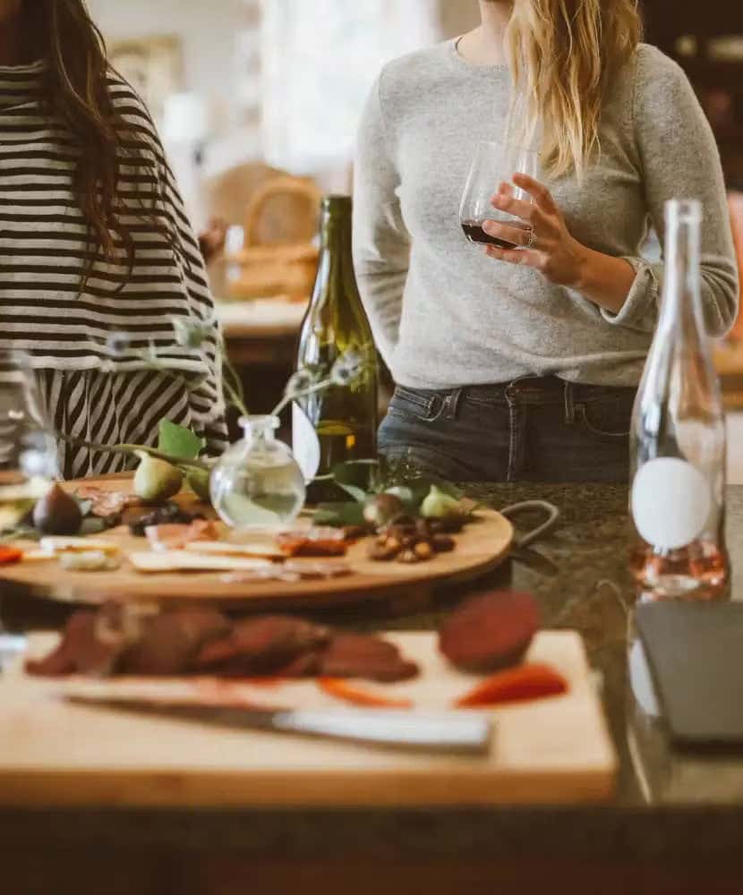 Two women standing at a kitchen counter with wine glasses, surrounded by bottles of wine and charcuterie boards filled with assorted meats, cheeses, and fruit.
