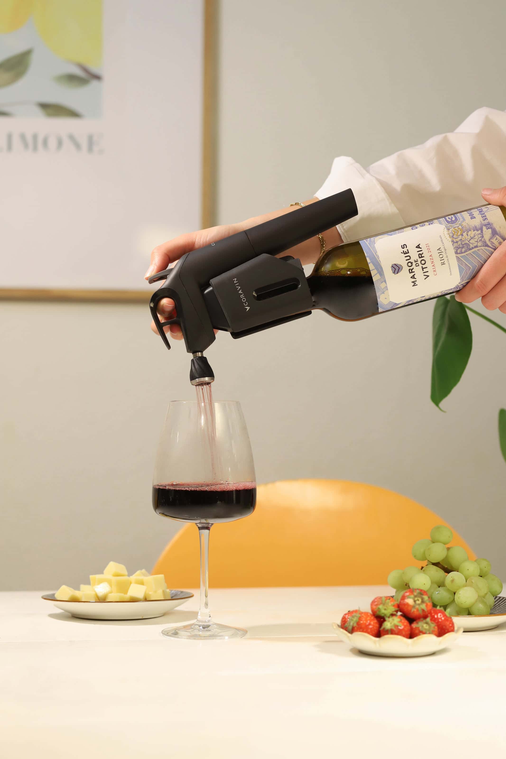 Close-up of a person using a Coravin wine preservation system to pour red wine from a bottle labeled 'Marqués de Vitoria' into a glass, with plates of cheese cubes, green grapes, and strawberries on a table in the foreground.