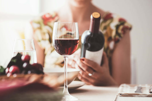Woman examining a bottle of red wine at a dining table, with a glass of wine and a bowl of fruit in the foreground.