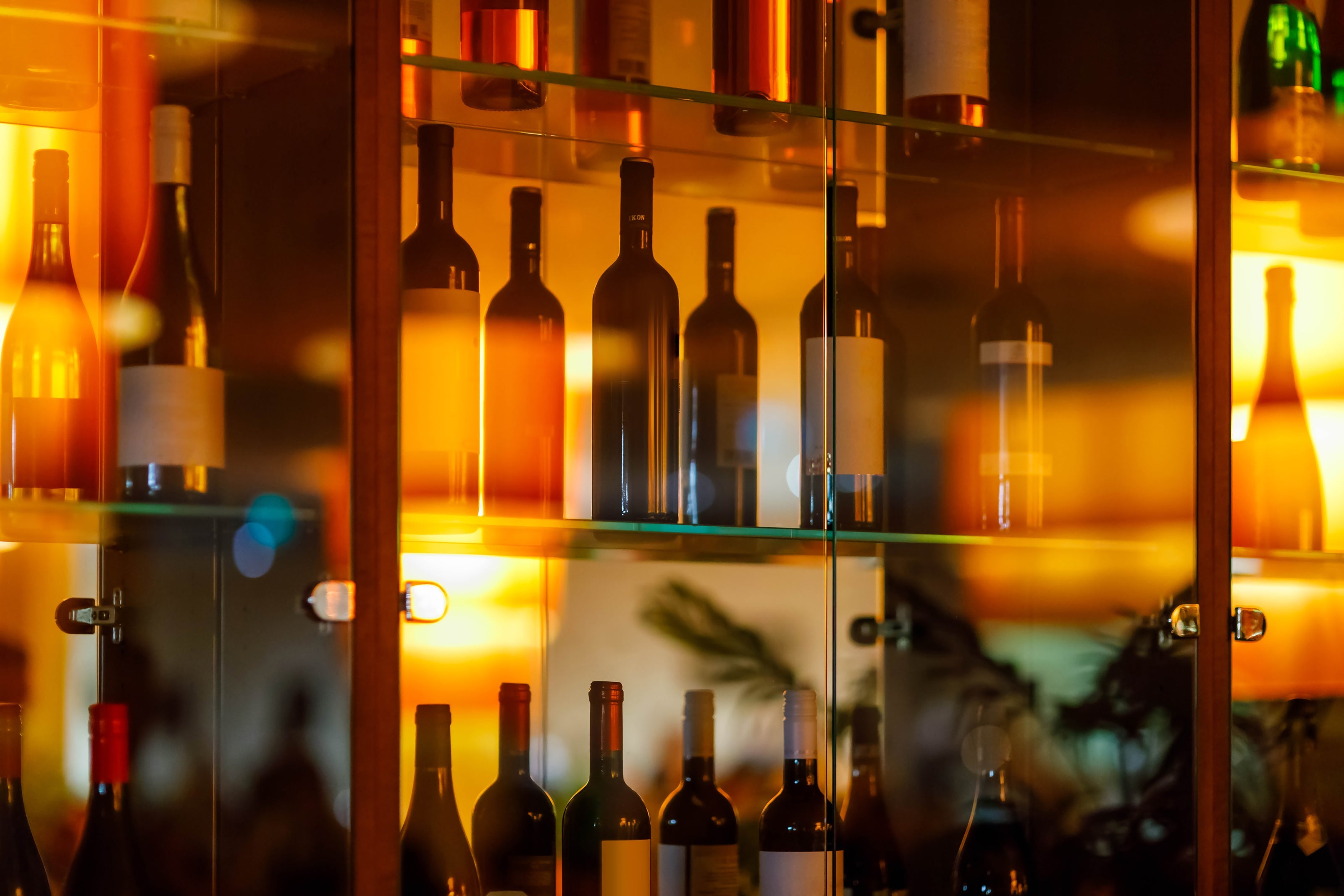 Wine bottles arranged on glass shelves behind a reflective glass cabinet, illuminated by warm ambient lighting in a cozy setting.