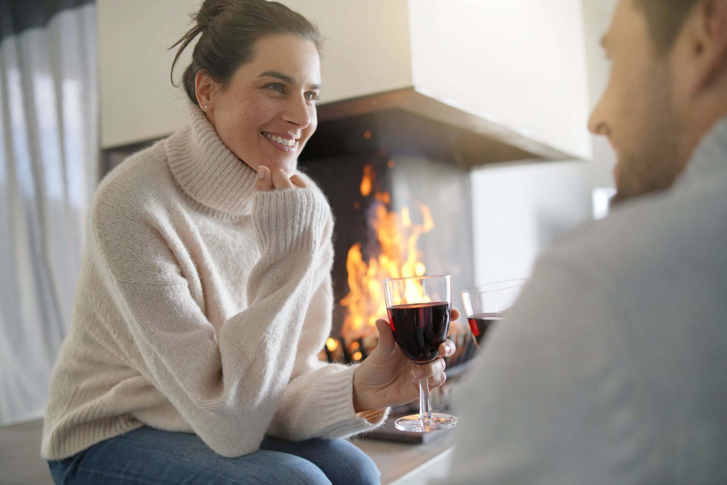 Smiling couple enjoying red wine by a warm fireplace, wearing winter sweaters and relaxing indoors.