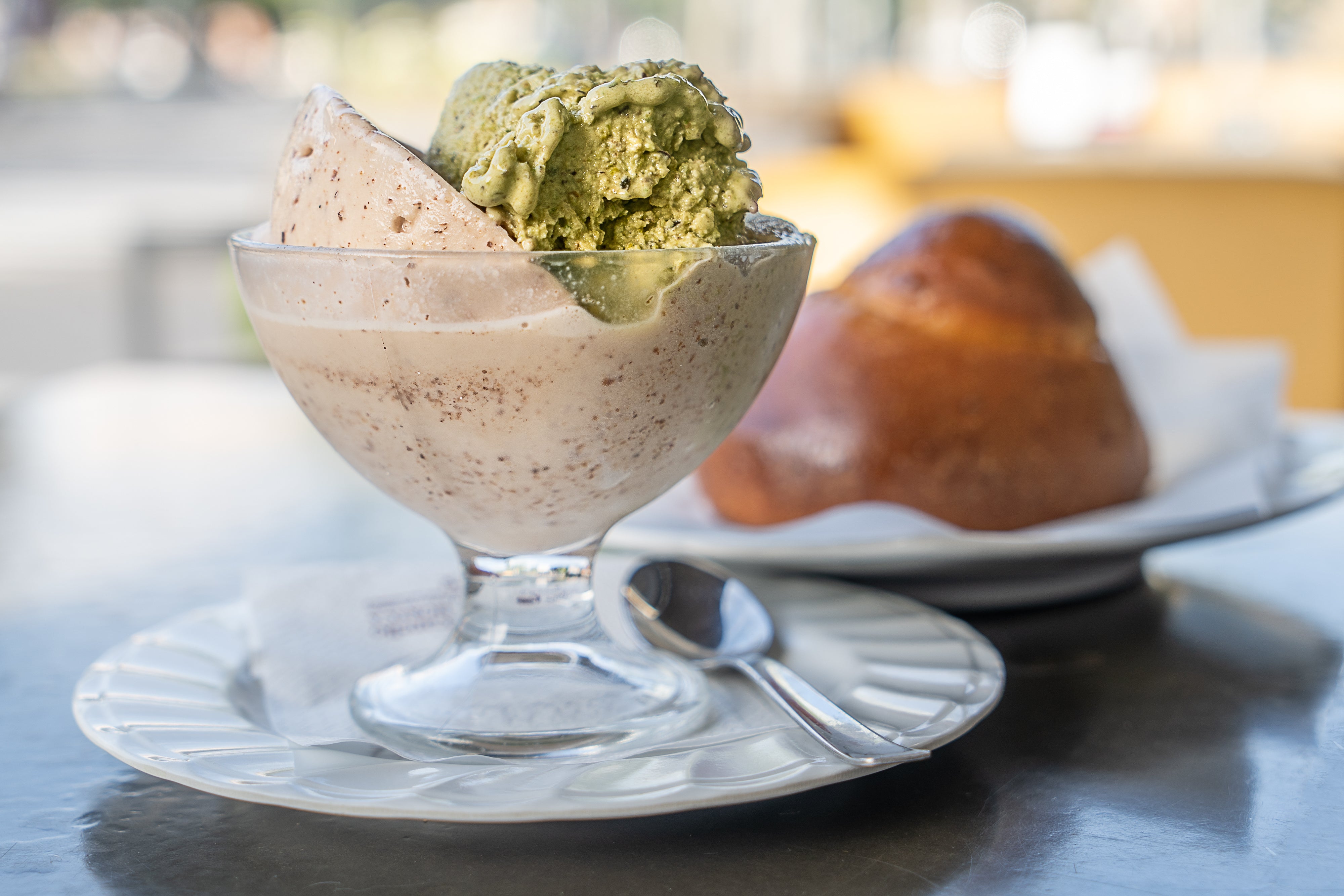 Traditional Sicilian granita with pistachio and chocolate flavors served in a glass bowl, accompanied by a soft brioche bun on a metal table.