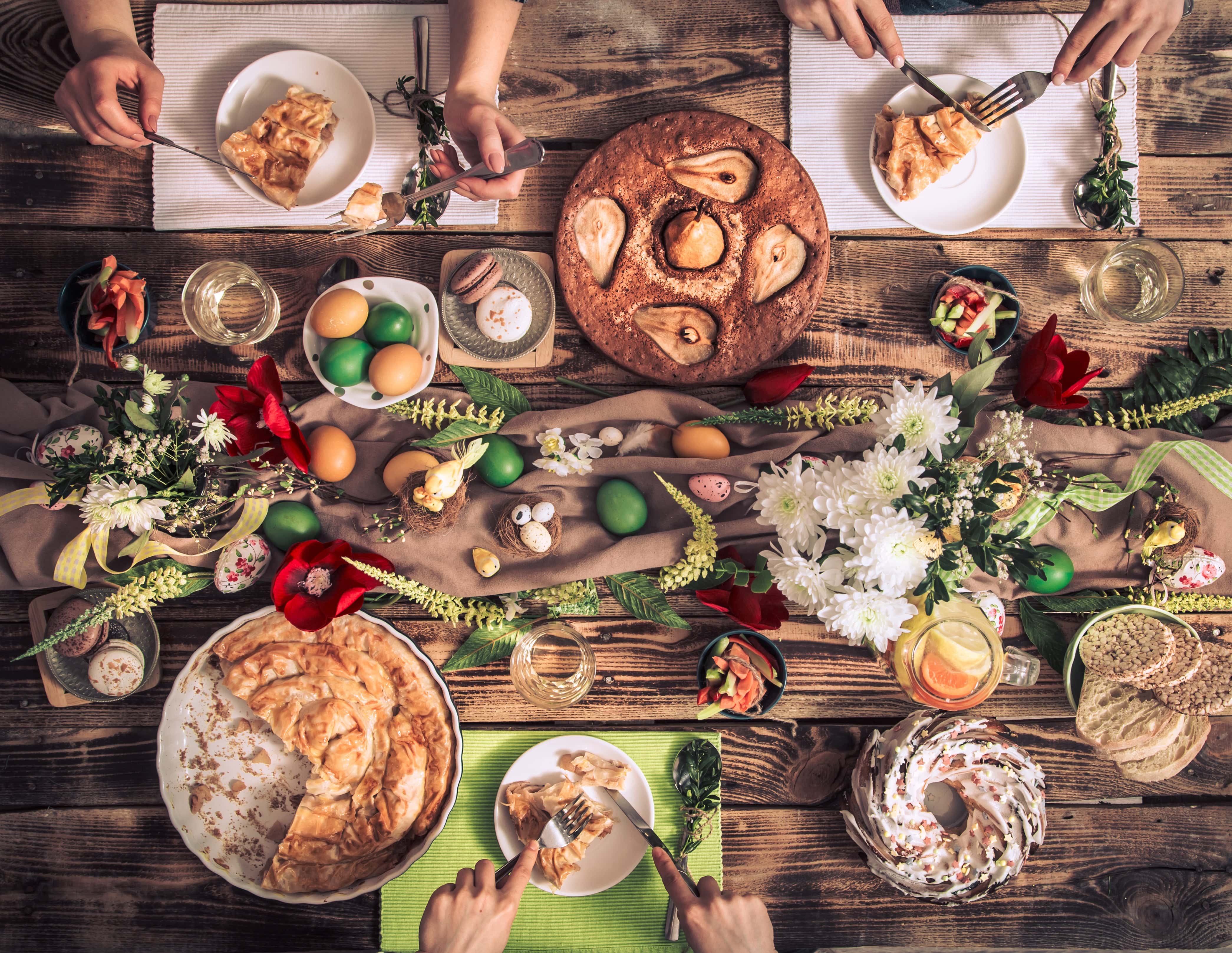 Overhead view of a festive table decorated with flowers, dyed Easter eggs, and traditional foods, with people enjoying slices of pie and glasses of white wine.