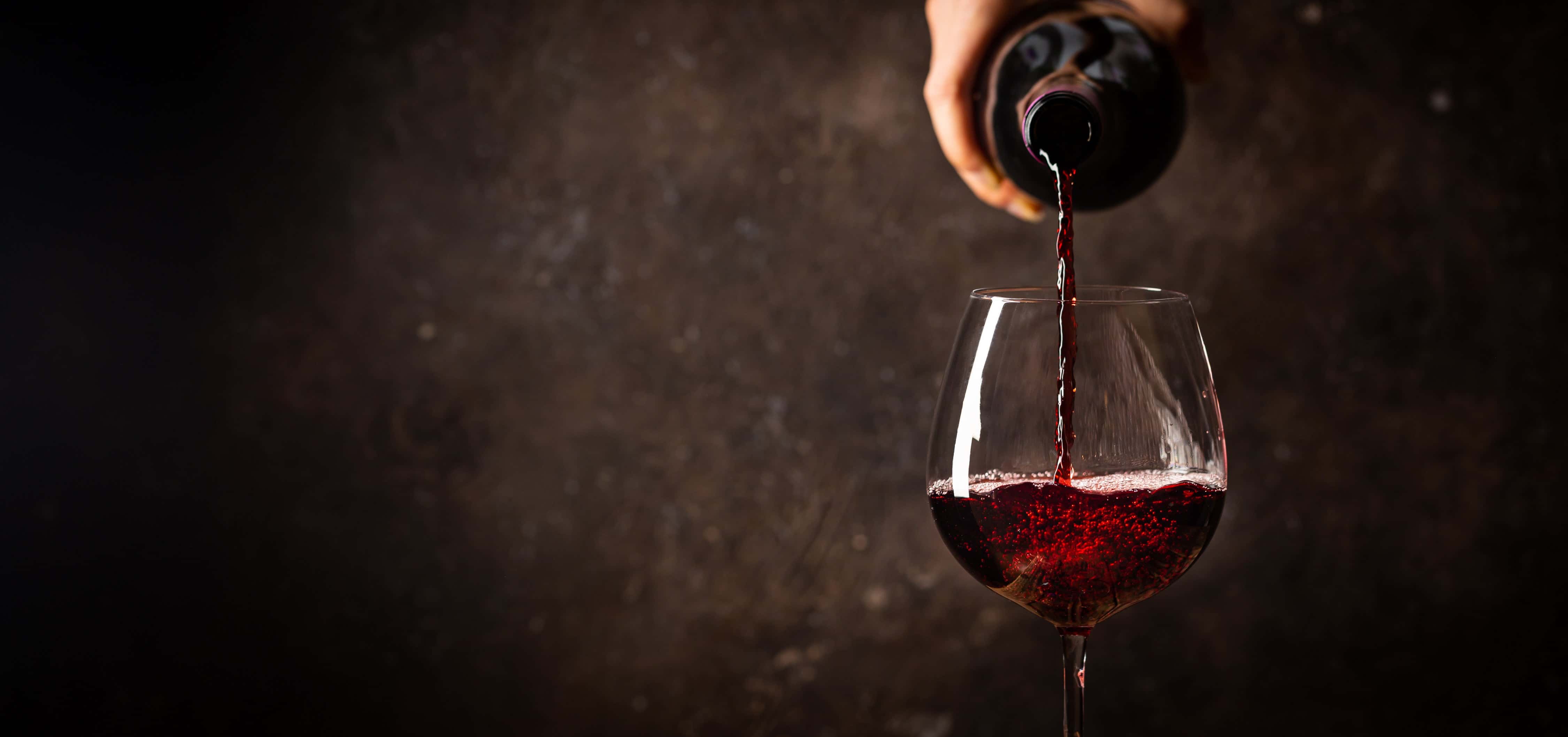 Red wine being poured into a clear wine glass against a dark textured background