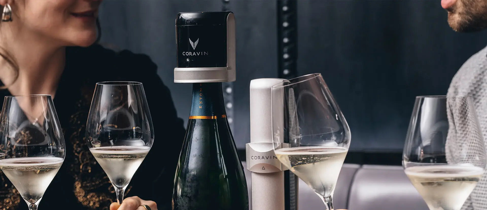 Two people enjoying glasses of sparkling wine at a table, with a bottle sealed using the Coravin Sparkling preservation system and the Coravin Sparkling Charger placed beside it.