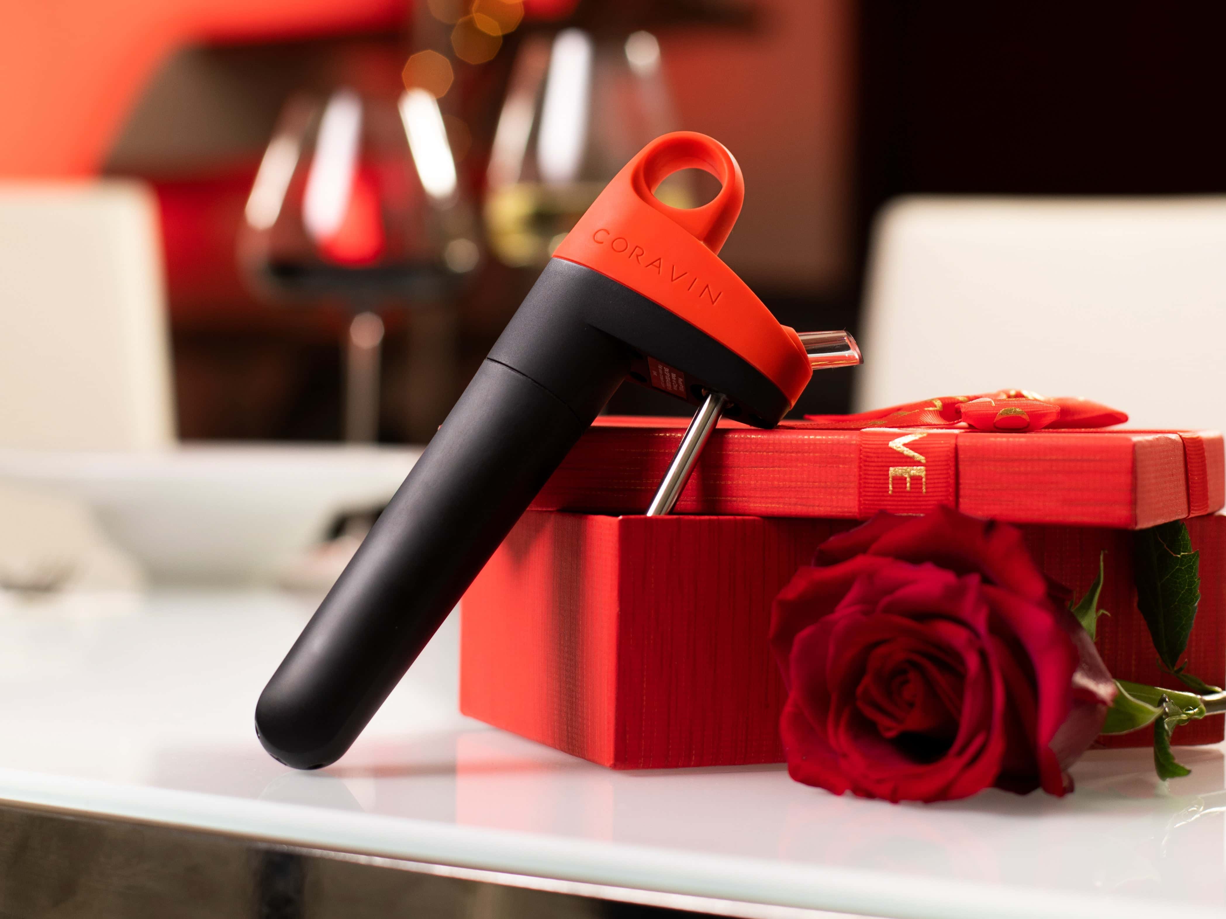 Coravin wine preservation system in black and red displayed on a red gift box with a rose, set on a romantic table with wine glasses in the background
