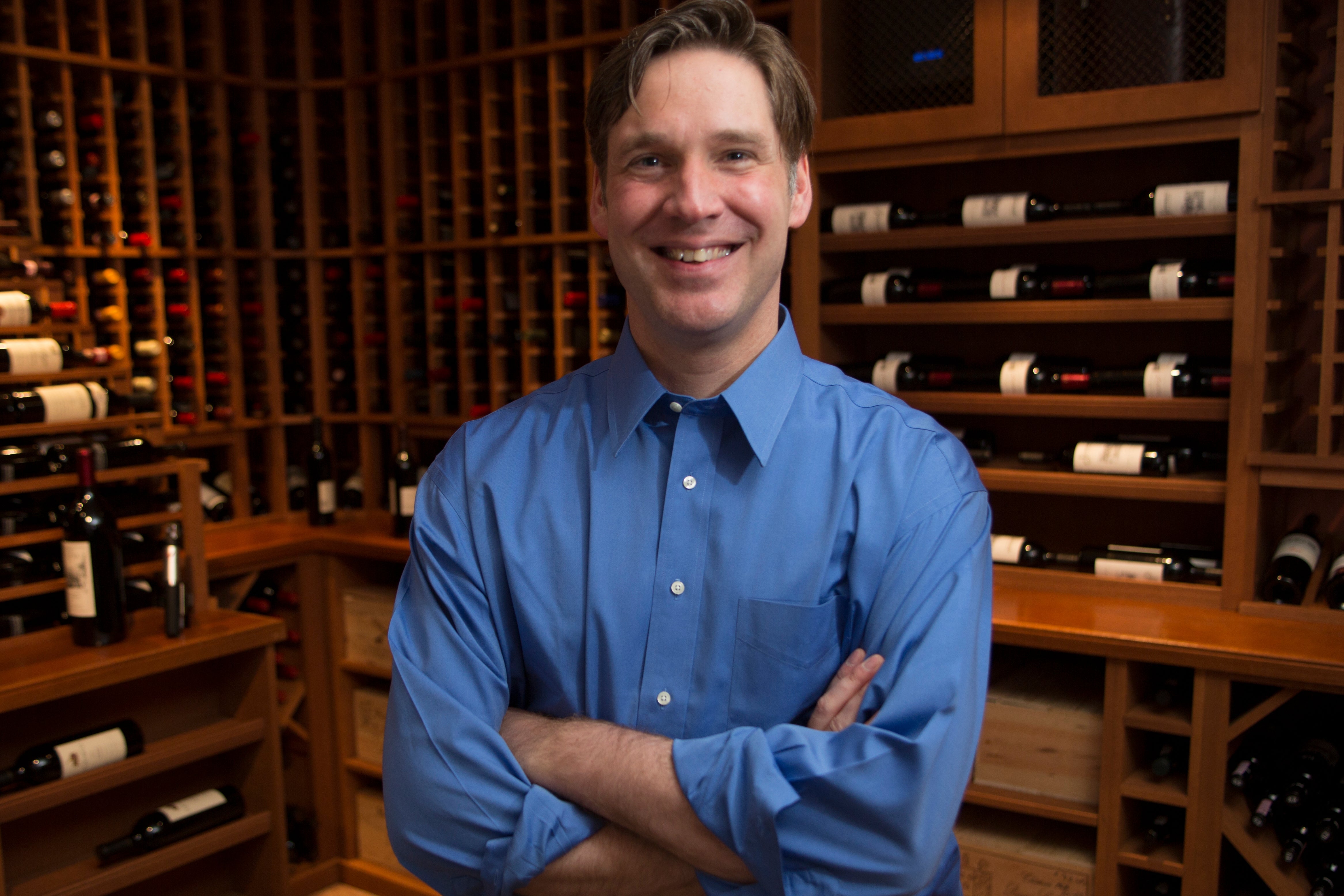 Man in blue shirt standing with arms crossed in a wine cellar filled with bottles.