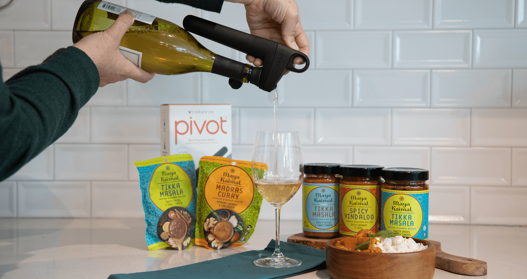 White wine being poured with a Coravin Pivot device into a glass, surrounded by Maya Kaimal Indian food products on a kitchen counter.