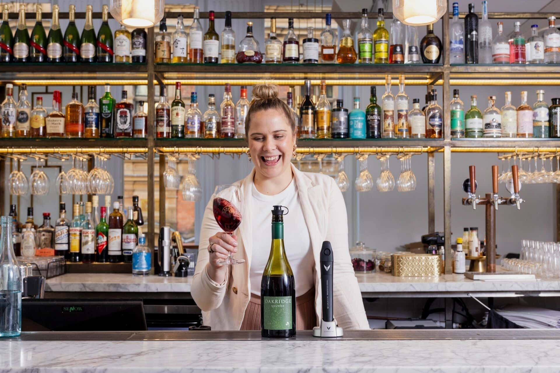 Smiling sommelier behind a marble bar holding a glass of red wine, with a Coravin wine preservation system and an open bottle of Oakridge wine in front of her, and a full back bar of liquor bottles and glasses behind.