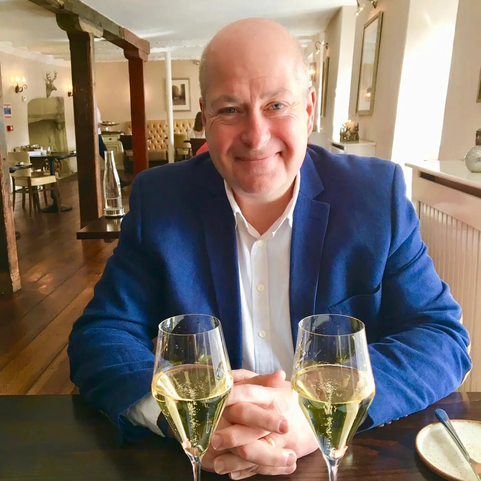 Smiling man in a blue blazer sitting at a table with two glasses of sparkling wine in a cozy restaurant.