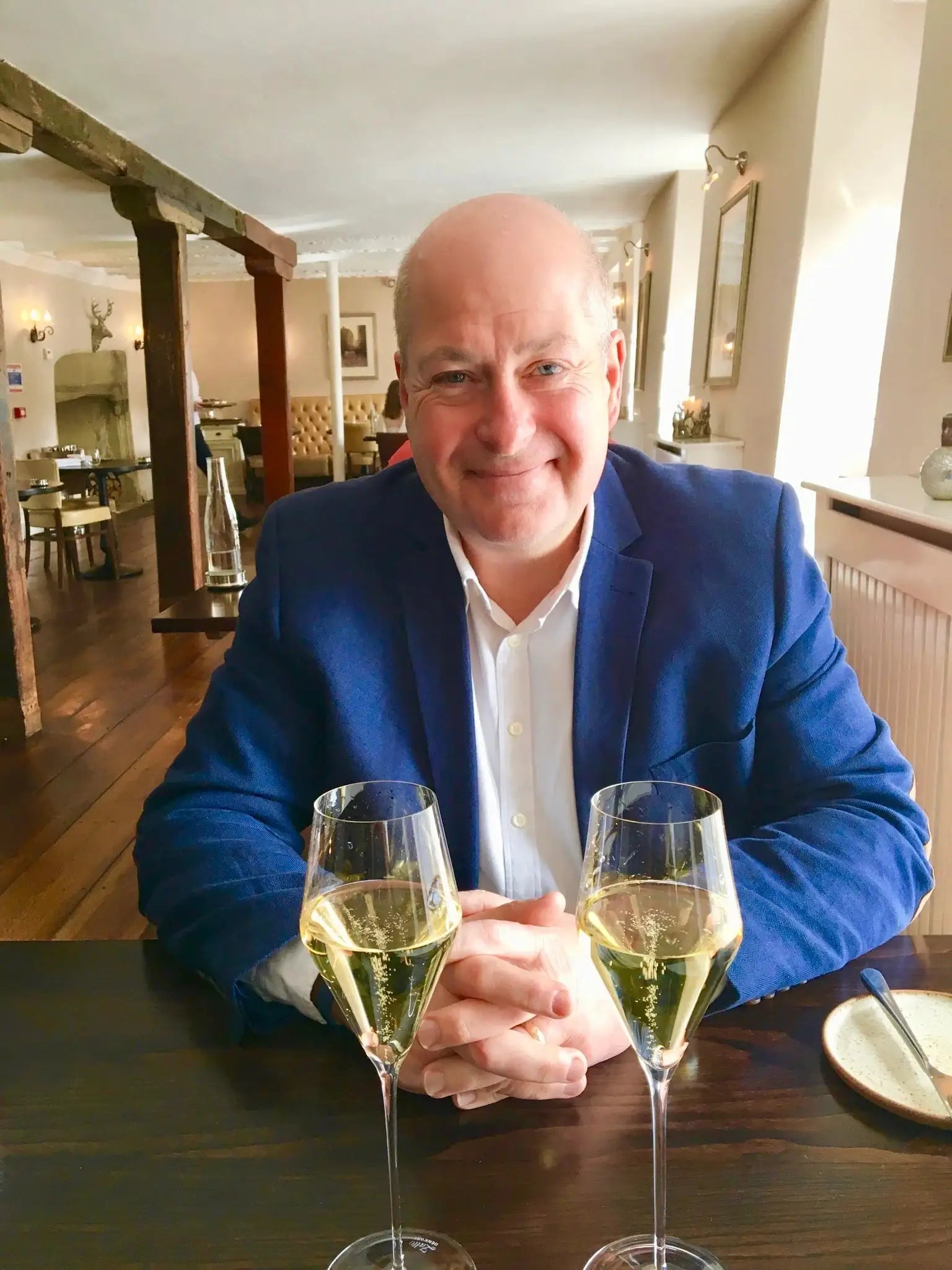 Smiling man in a blue blazer sitting at a table with two glasses of sparkling wine in a cozy restaurant.