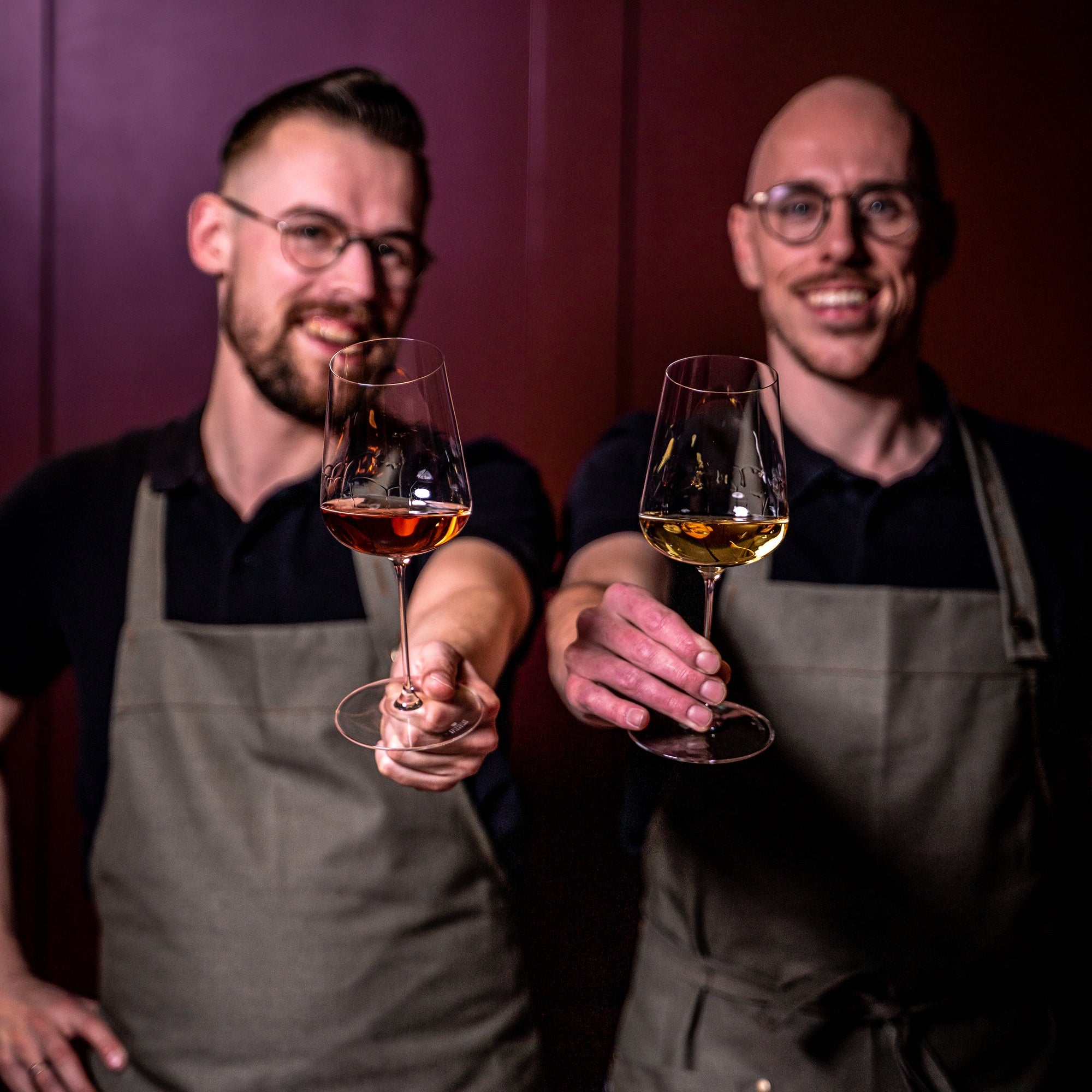 Two smiling bartenders in aprons holding glasses of wine toward the camera.