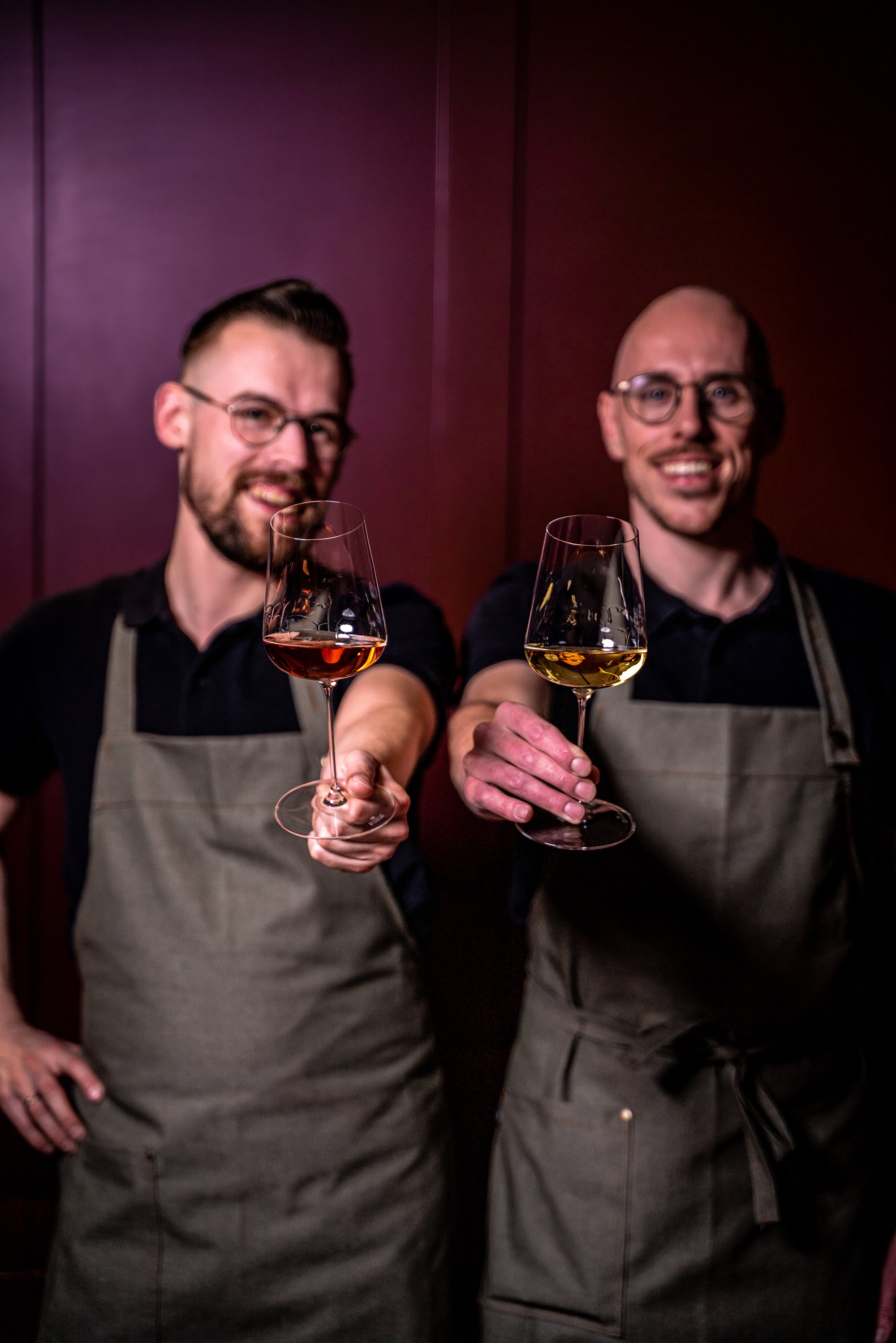 Two smiling bartenders in aprons holding glasses of wine toward the camera.