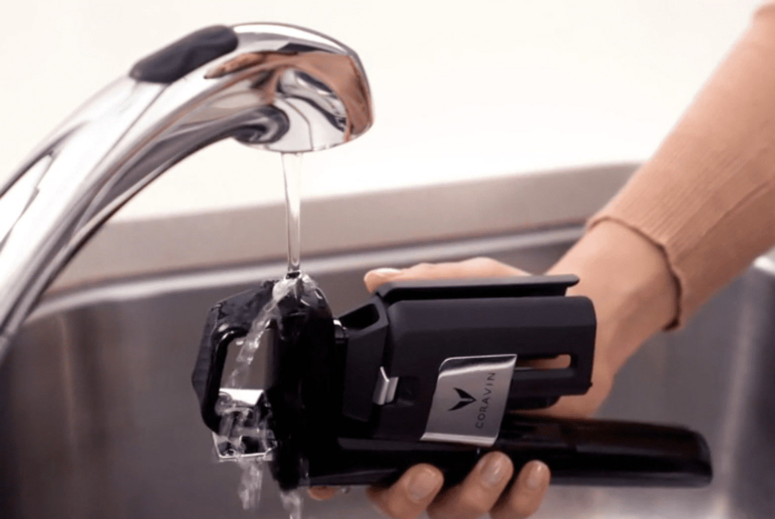Person rinsing a Coravin wine preservation system under running water from a kitchen faucet over a sink.