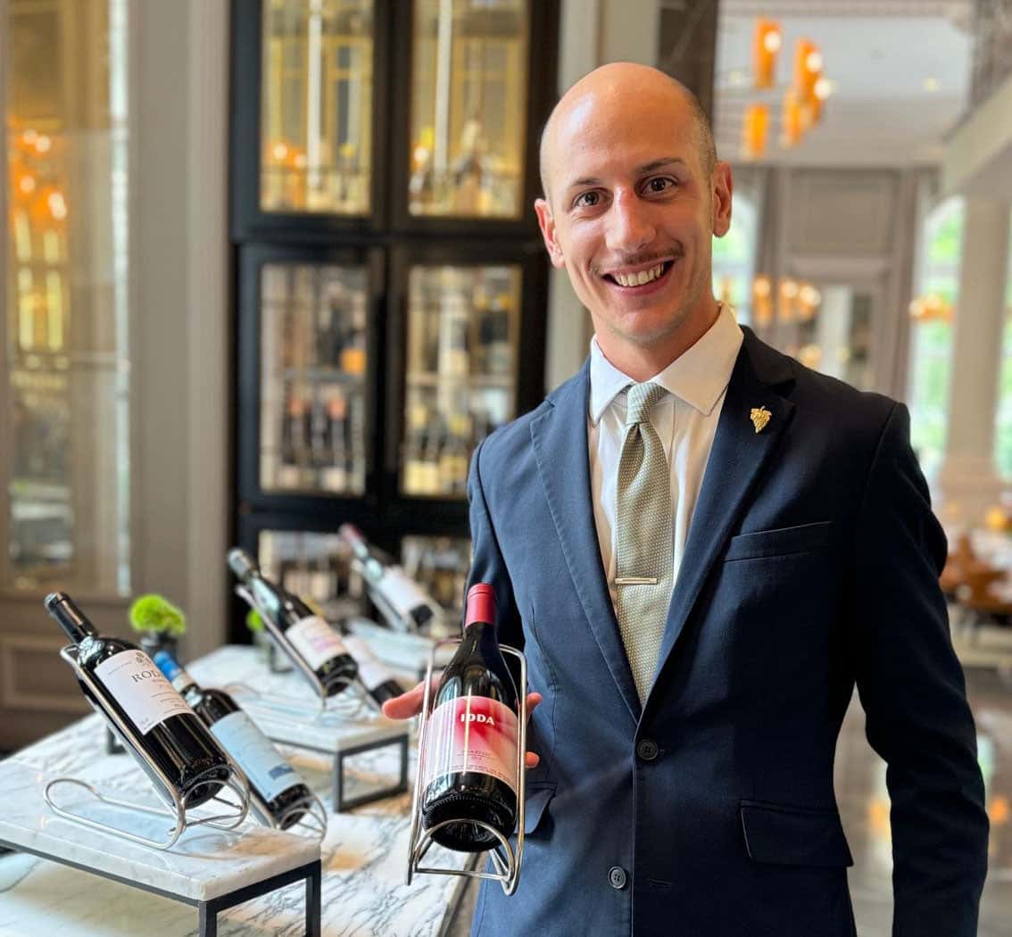 Smiling sommelier in a suit holding a bottle of red wine in an elegant wine tasting room.