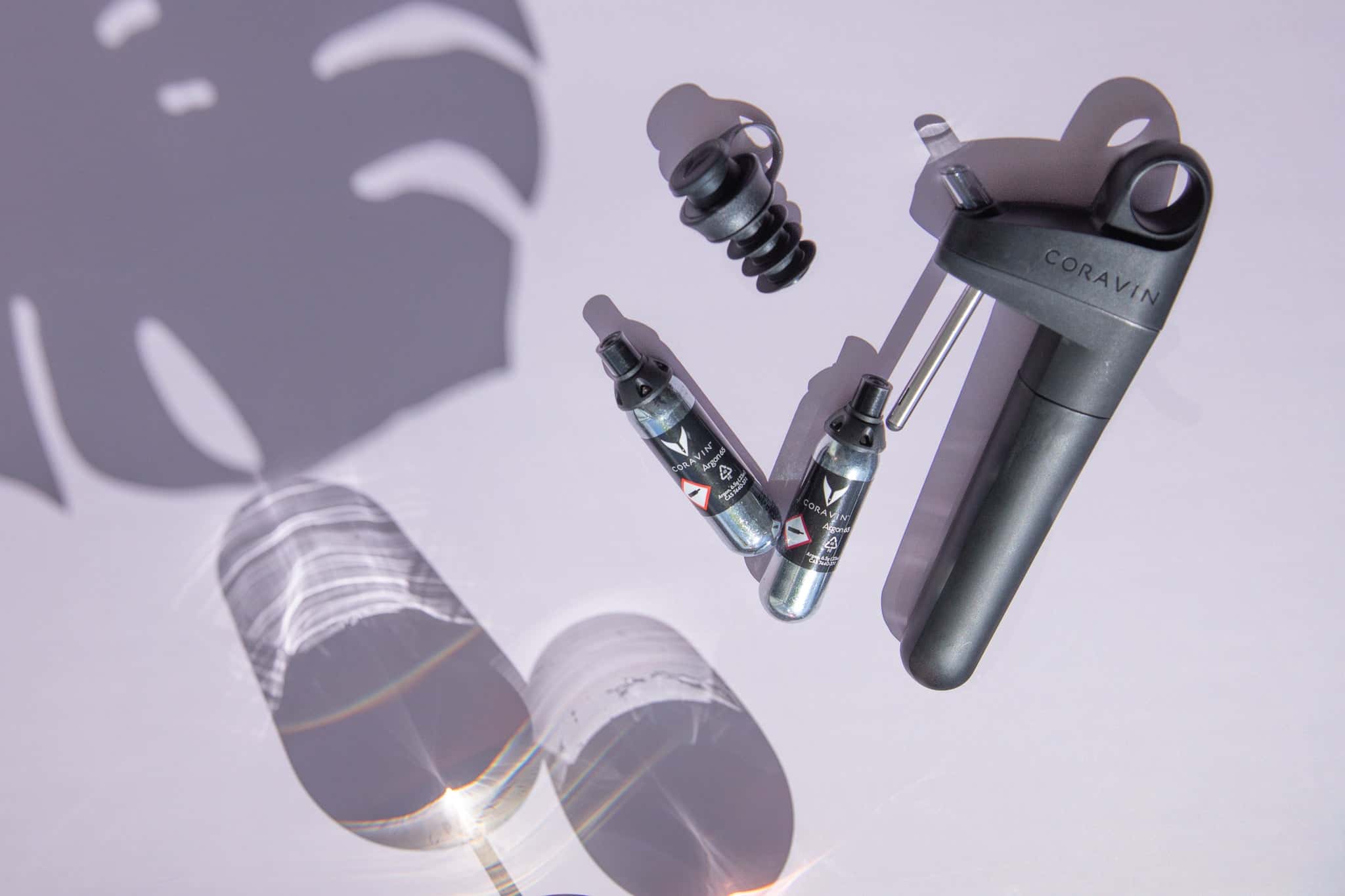 Coravin Pivot wine preservation system with argon capsules, stopper, and glass reflections on a pastel background with tropical leaf shadow