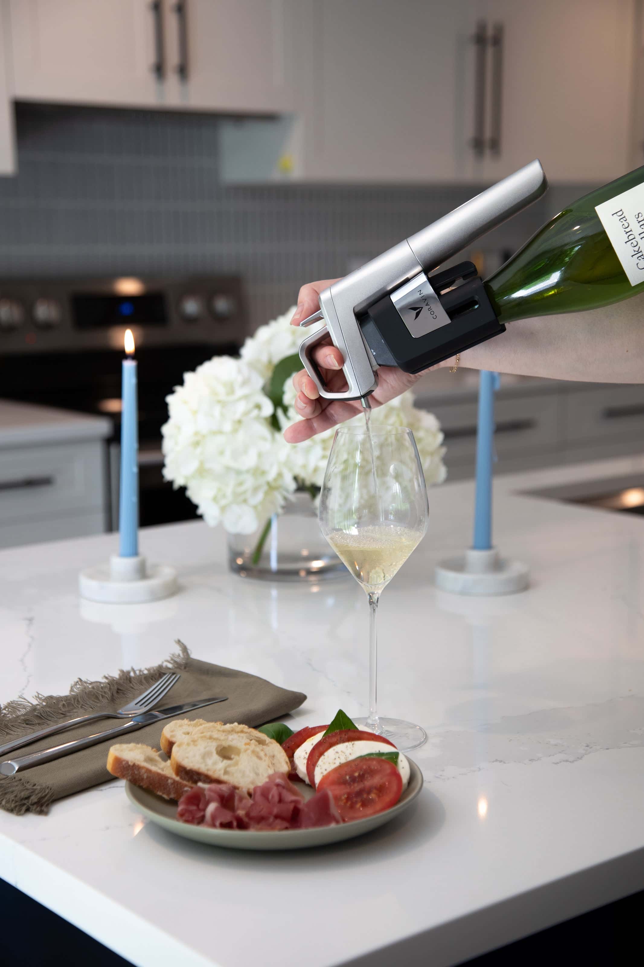 Pouring white wine using a Coravin Timeless Six preservation system into a glass on a modern kitchen counter, with a plate of prosciutto, mozzarella, tomato, and bread.