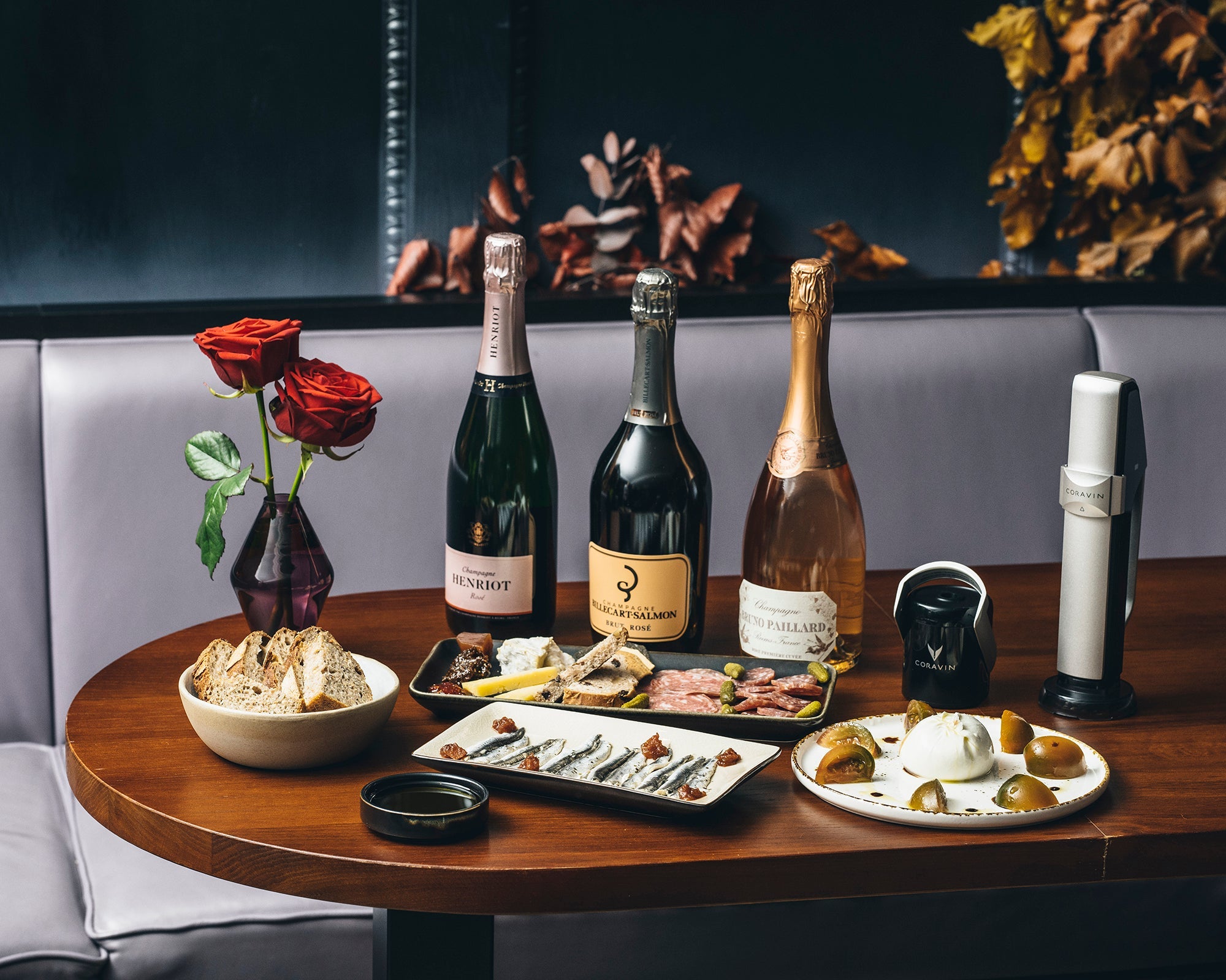 Table set with Coravin Sparkling wine preservation system, three bottles of champagne, gourmet charcuterie, bread, cheese, and a vase with red roses.