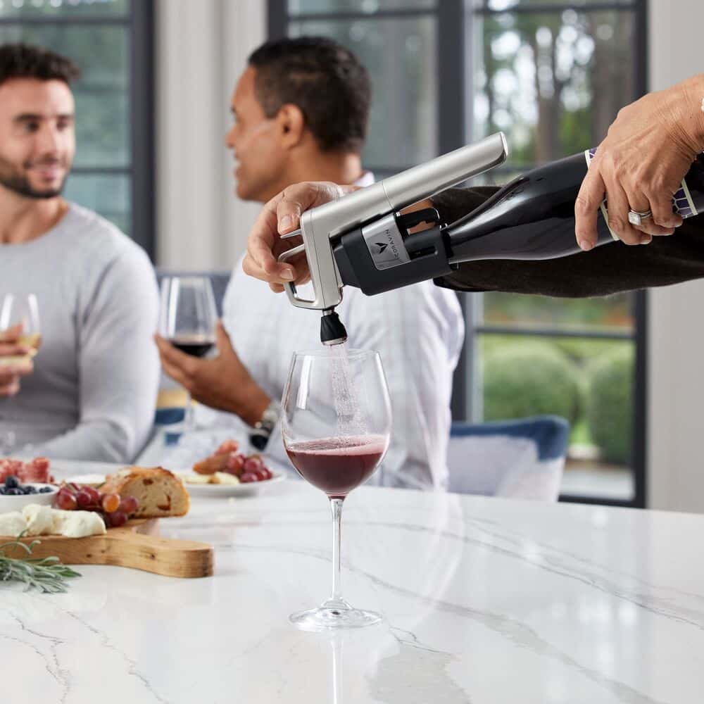Person using a Coravin wine preservation system to pour red wine at a social gathering, with guests enjoying charcuterie and wine around a marble countertop.