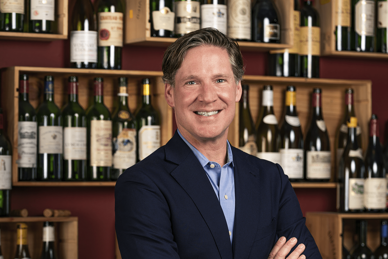 Smiling man in a navy suit standing in front of a wine wall with assorted wine bottles.