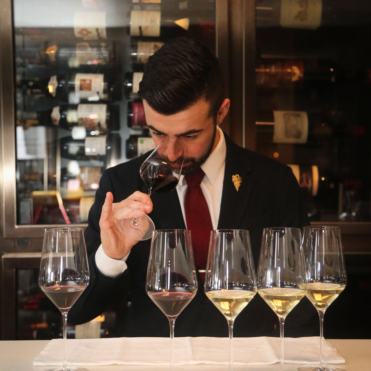 Sommelier in suit tasting red wine with a lineup of wine glasses in front of a wine cellar.