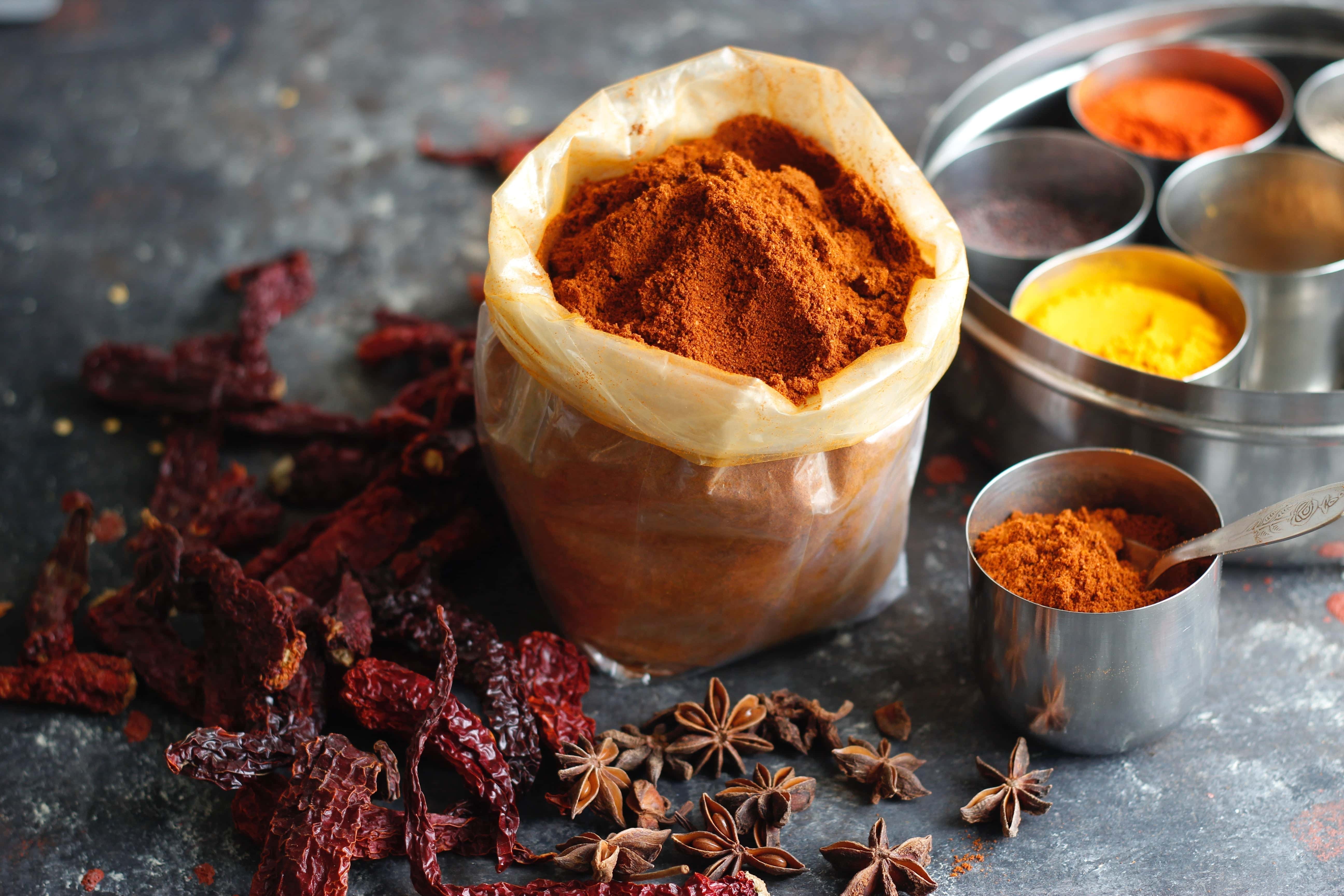 Assorted Indian spices including dried red chilies, star anise, and colorful ground powders in metal containers on a dark rustic background.