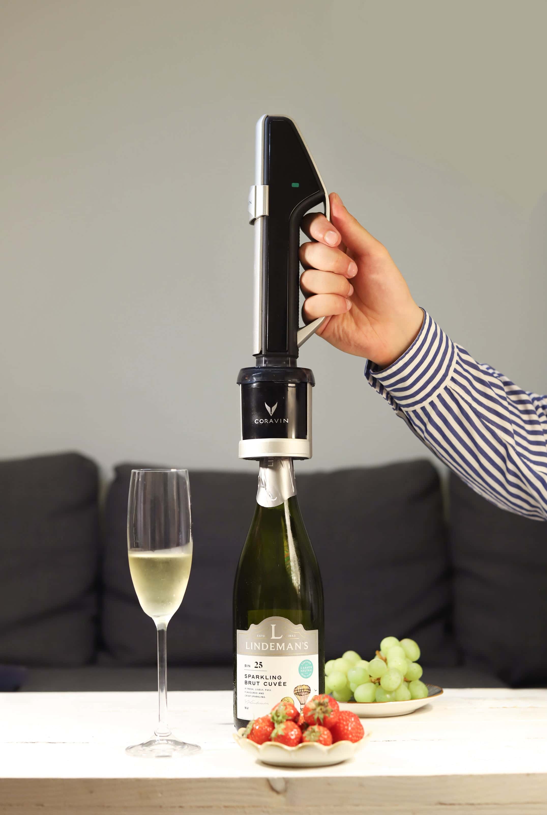 Person using Coravin Sparkling System to pour Lindeman’s Brut Cuvée into a flute glass, with strawberries and green grapes on a table.