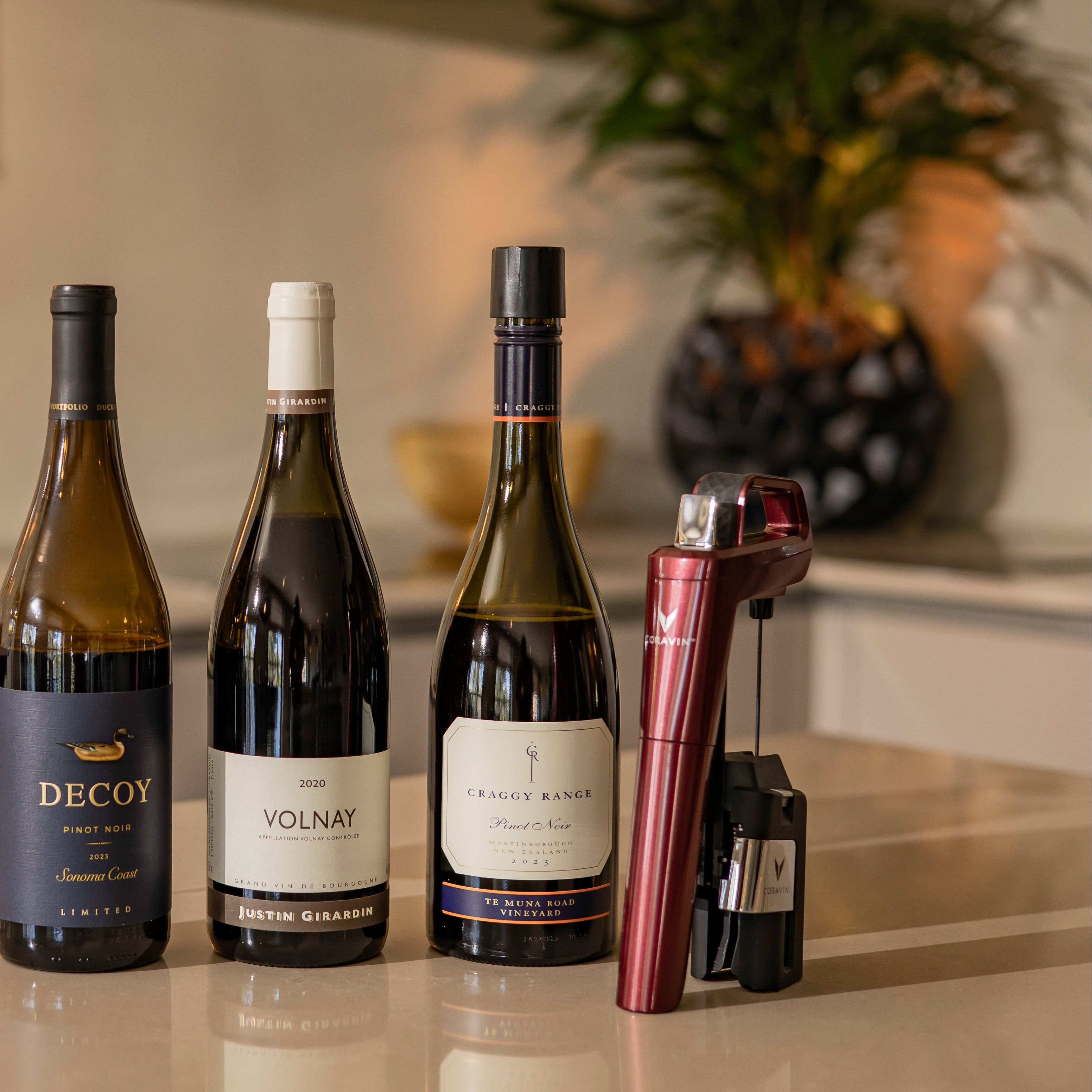 Four wine bottles and a coravin red wine preservation system on a reflective surface with a blurred background