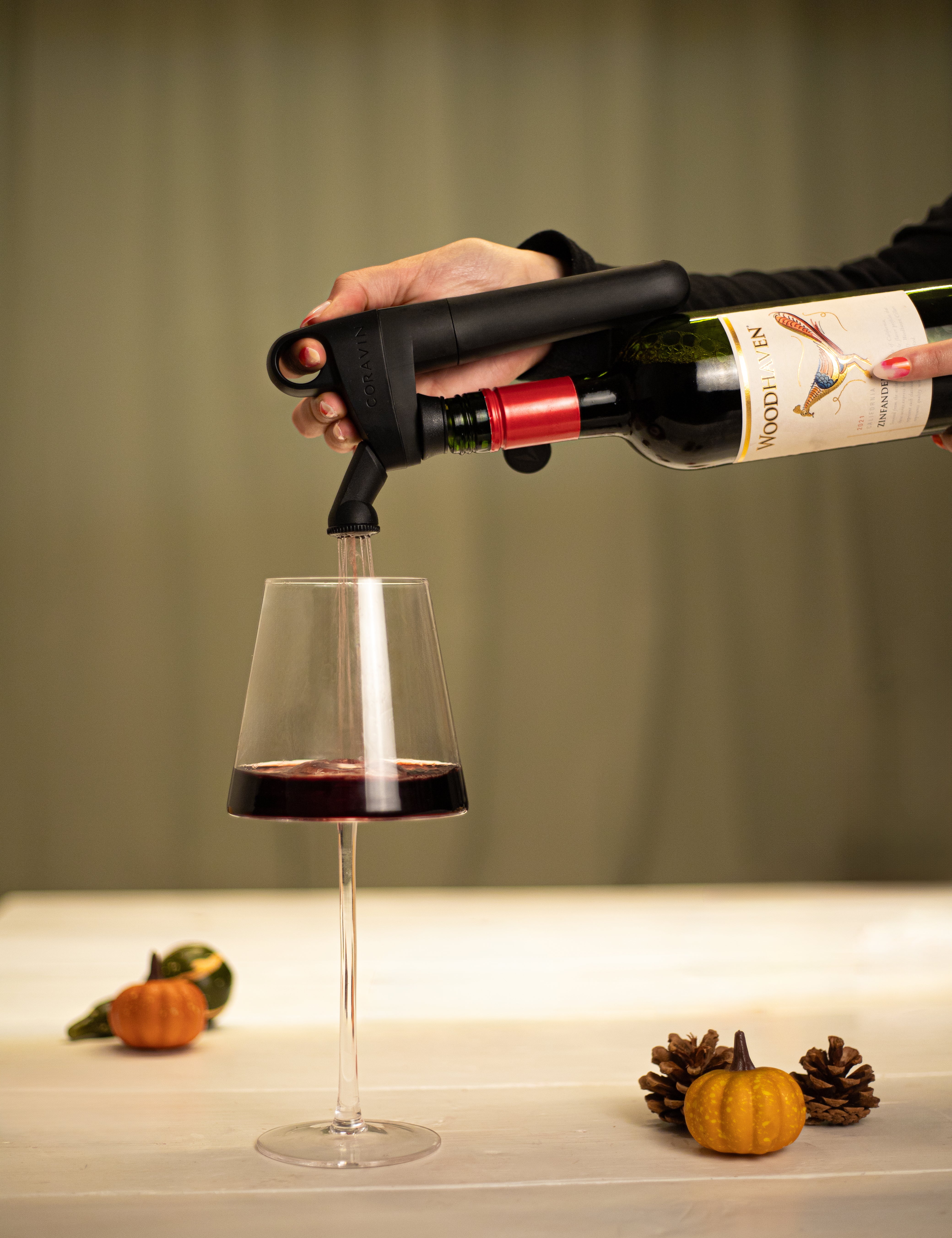 A hand uses a Coravin wine opener to pour red wine into a glass, with pumpkins and pine cones on the table.